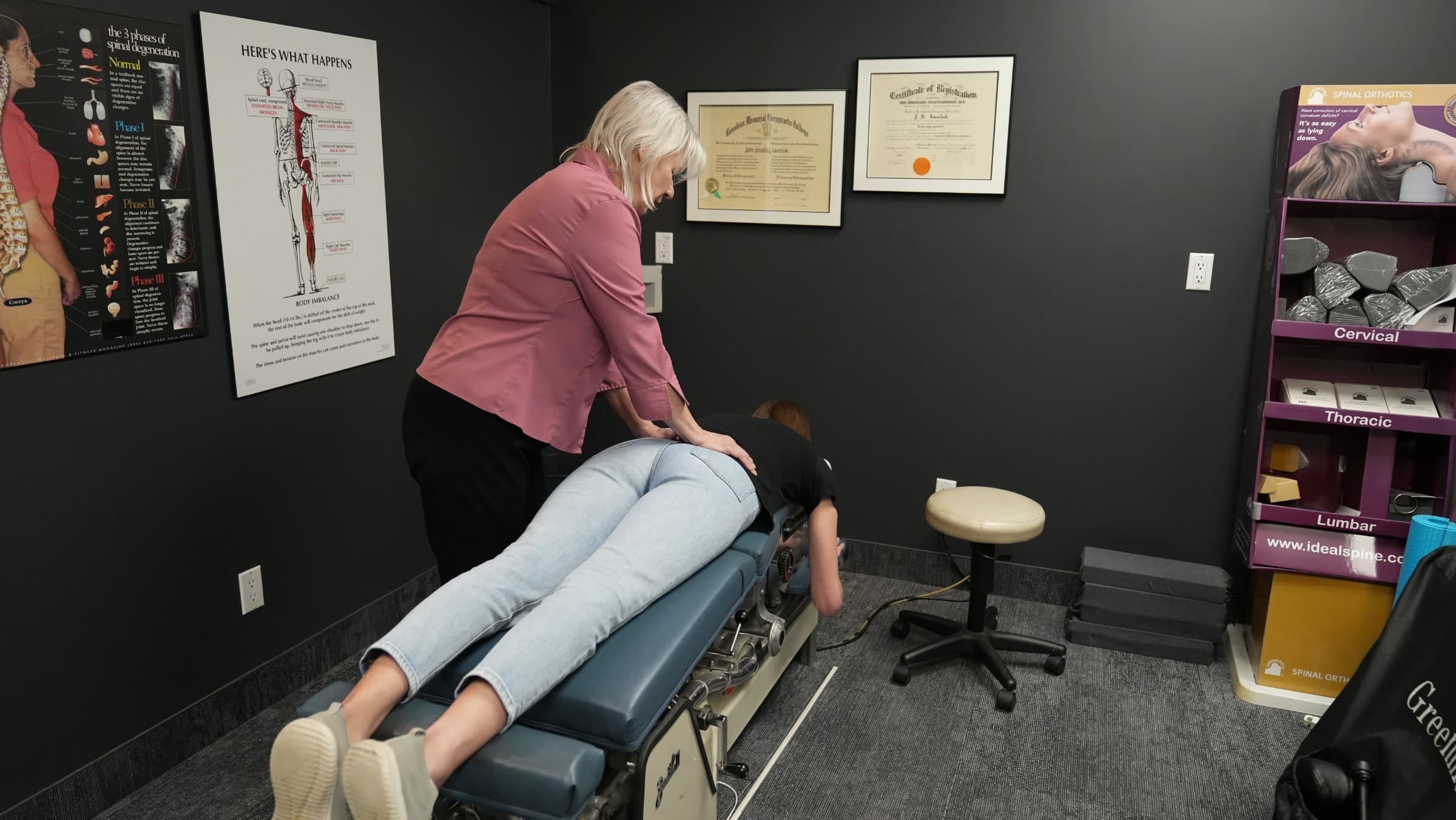 A chiropractor in a pink shirt performs a spinal adjustment on a patient lying face-down on a treatment table in a Burlington chiropractic clinic, with anatomical charts and certification documents visible on the dark walls