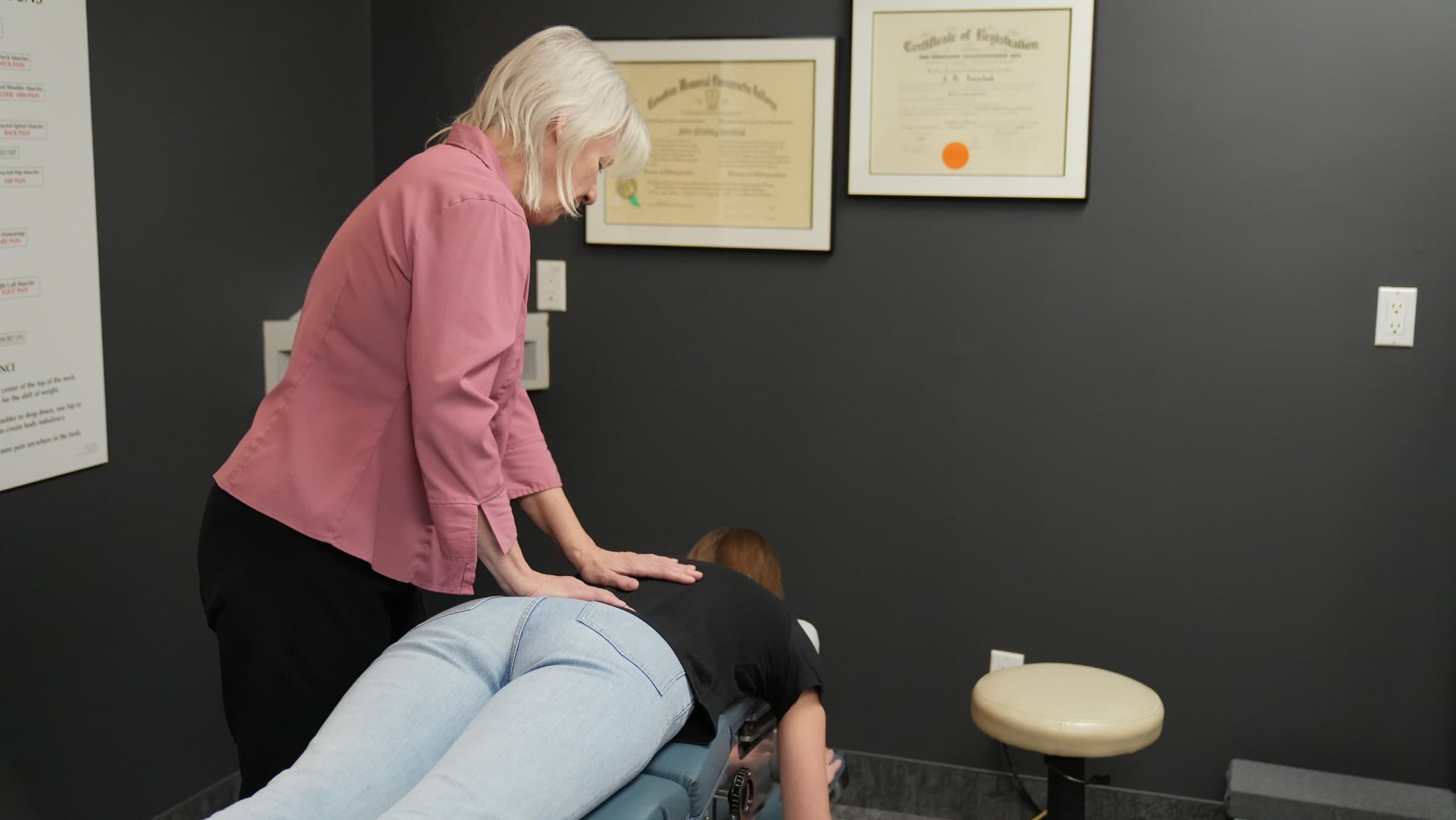 A chiropractor in a pink shirt performs a back adjustment on a patient in a treatment room at Life Chiropractic Centre in Burlington, Ontario, with framed certificates displayed on the dark gray wall