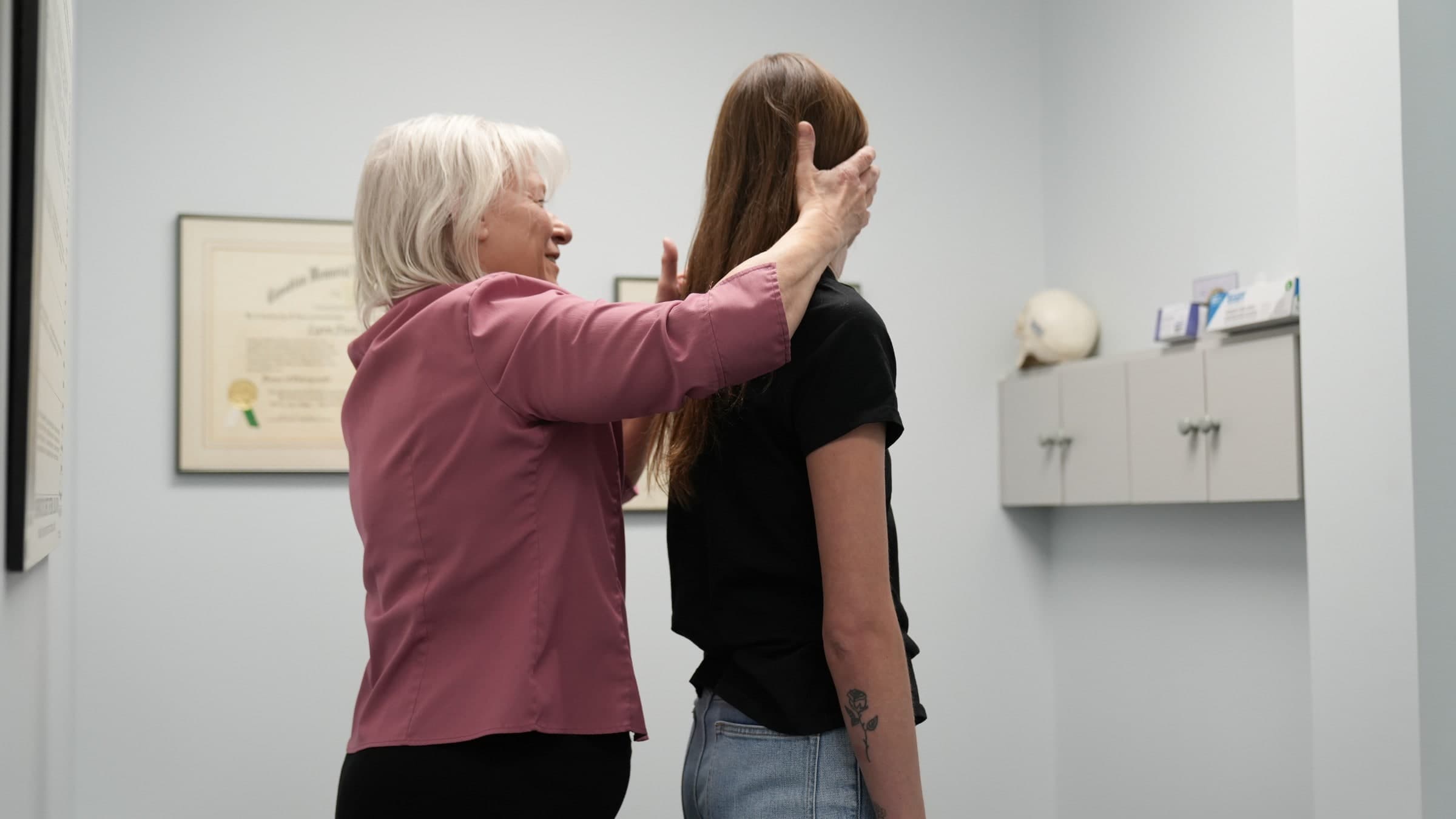 A chiropractor in a maroon shirt performs a neck examination on a patient with brown hair in a clinical treatment room in Burlington, Ontario
