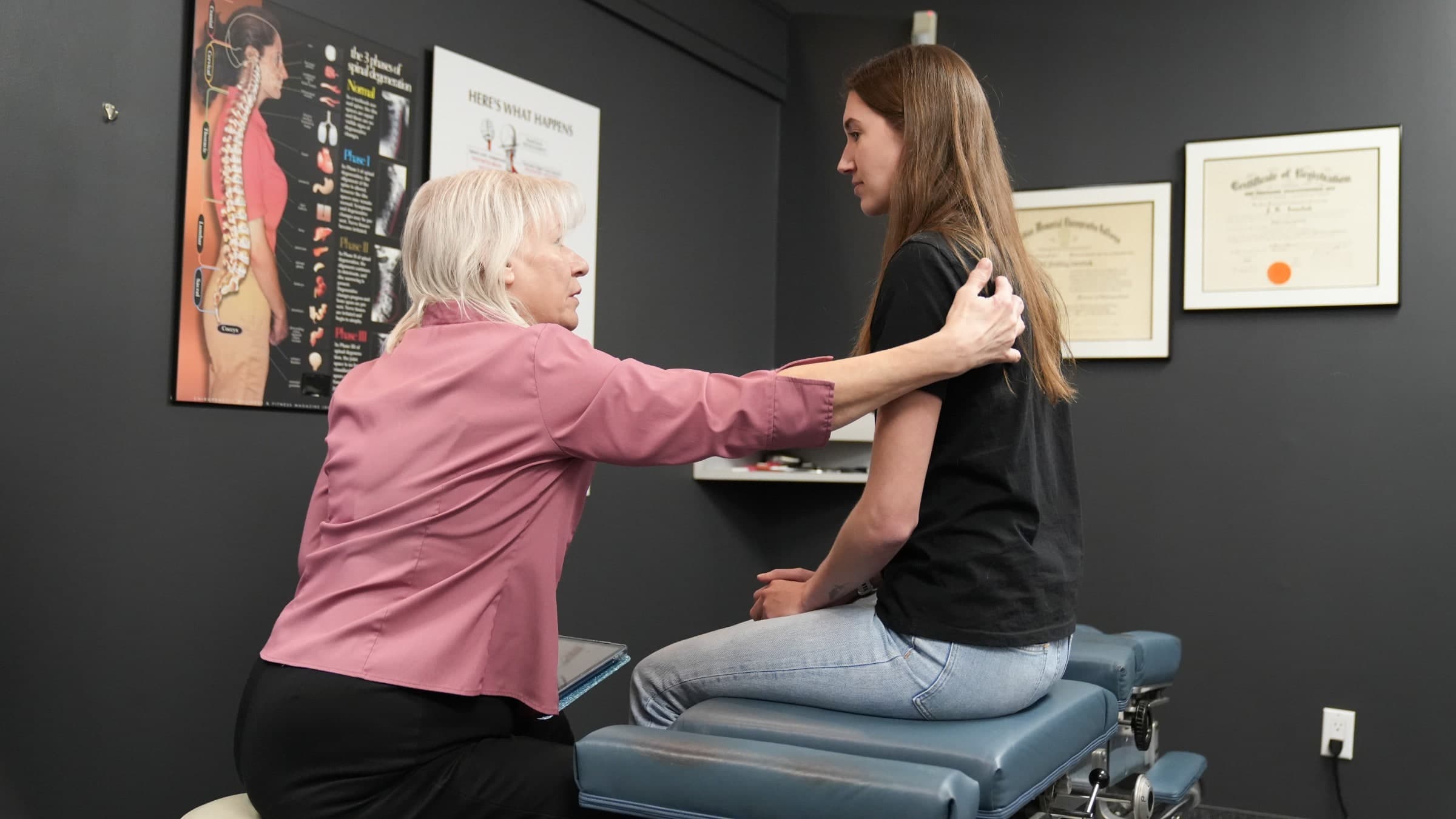 A chiropractor in a pink jacket examines a patient's shoulder while the patient sits on an examination table in a clinical room in Burlington, with anatomical charts on the wall behind them