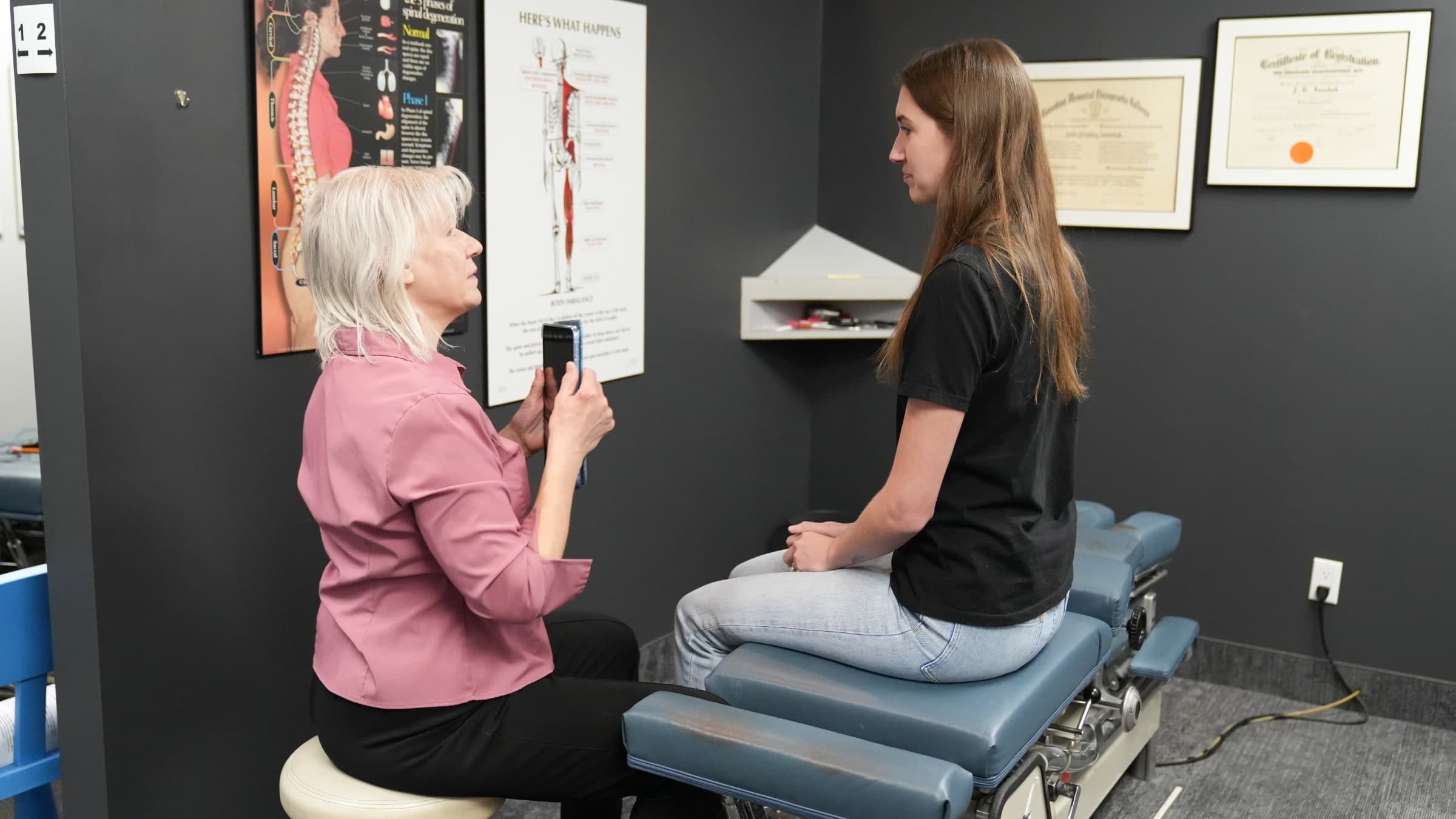 A chiropractor in a pink jacket records a video on her phone while speaking with a patient sitting on an examination table in a clinical room at Life Chiropractic Centre in Burlington, Ontario