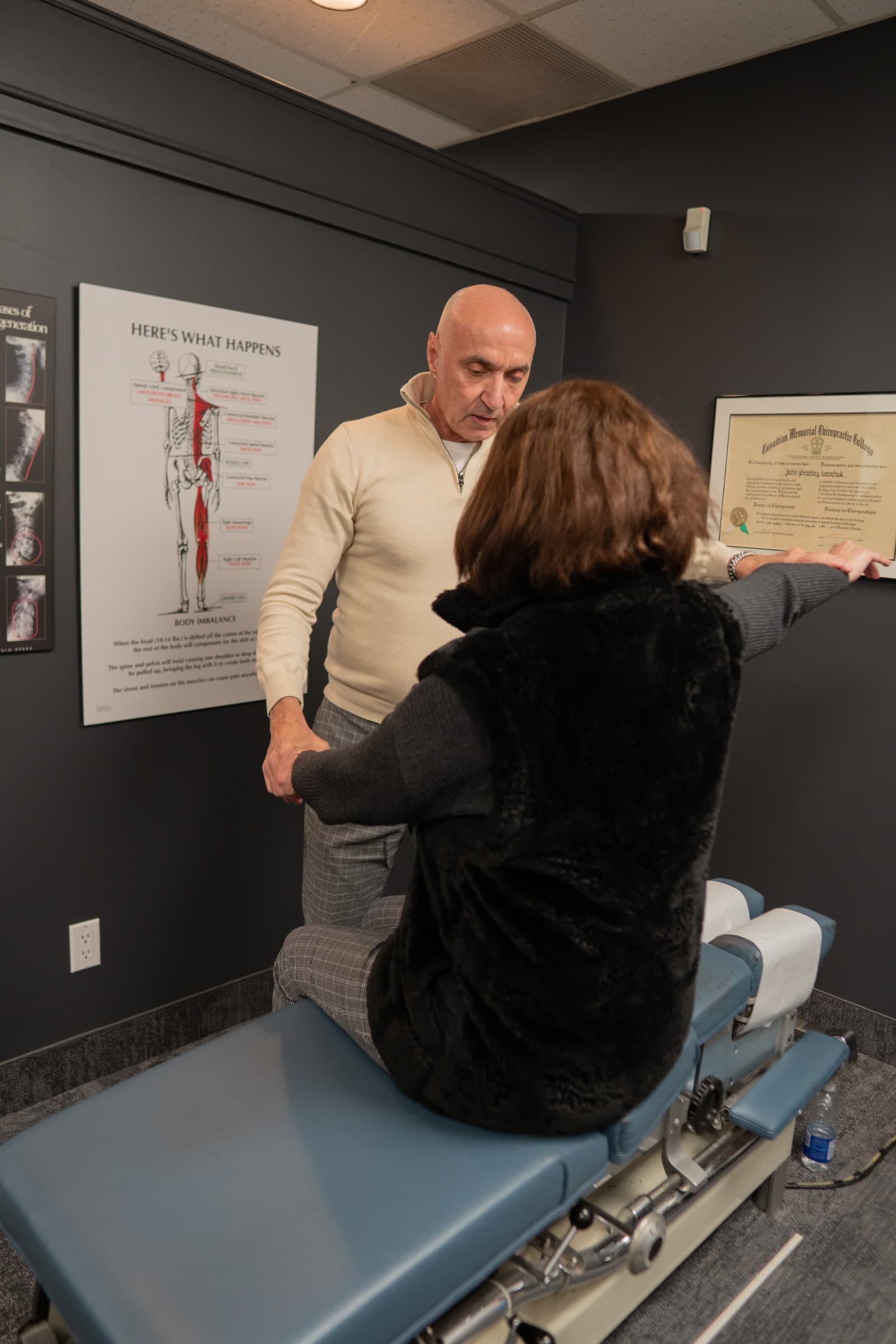 A chiropractor performs a spinal examination on a patient seated on a treatment table in a clinical room at Life Chiropractic Centre in Burlington, Ontario, with anatomical diagrams displayed on the dark walls behind them