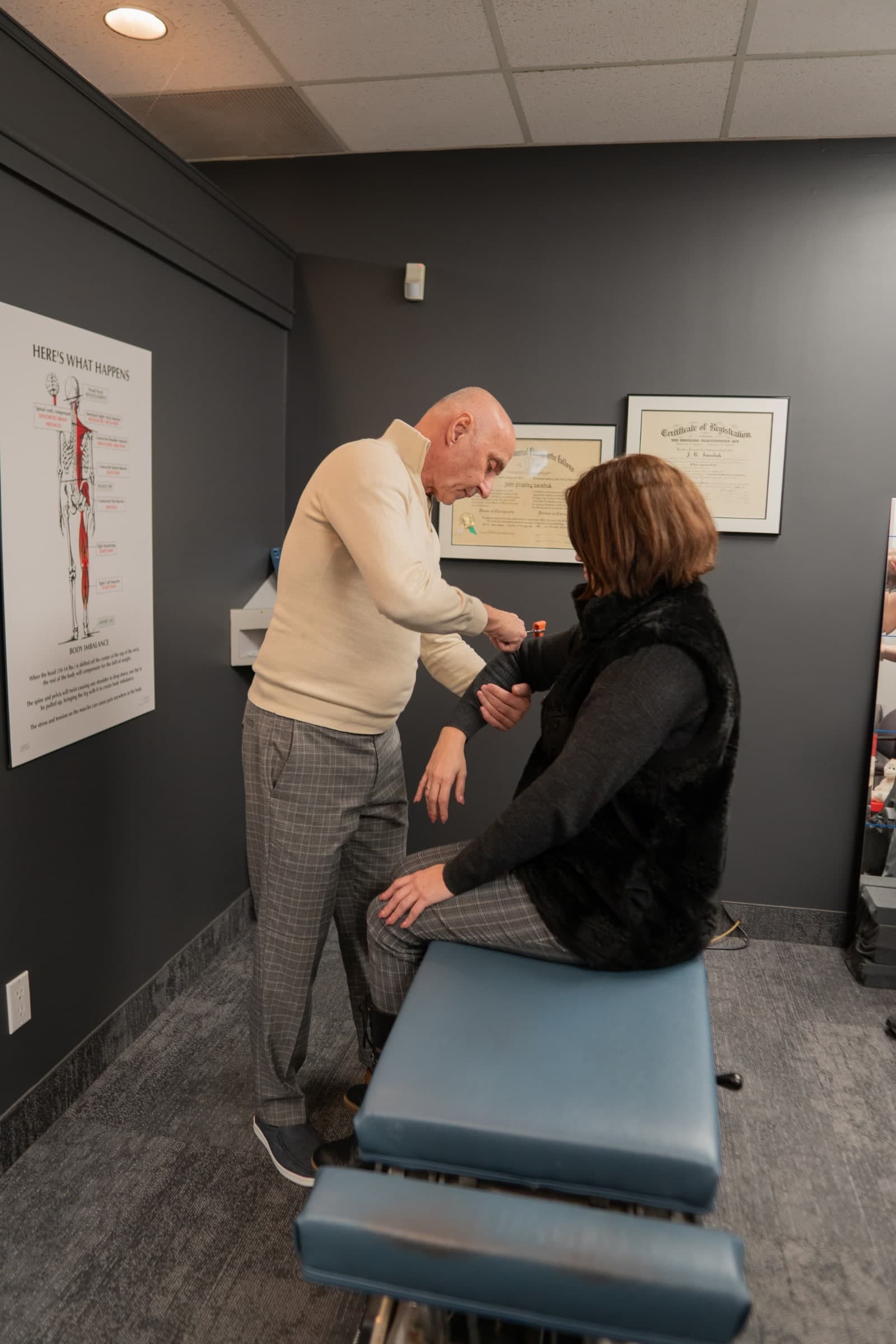 A chiropractor performs an examination on a patient's arm in a treatment room at Life Chiropractic Centre in Burlington, Ontario, with anatomical diagrams displayed on the dark gray walls
