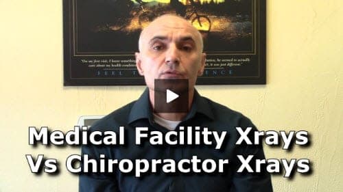 A man in dark clothing speaks directly to the camera with text overlay reading "Medical Facility Xrays Vs Chiropractor Xrays" in front of a framed poster on a yellow wall